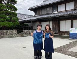 Two women holding newly indigo-dyed cloth pieces standing in the courtyard in front of the traditional wooden Okumura Family Residence.