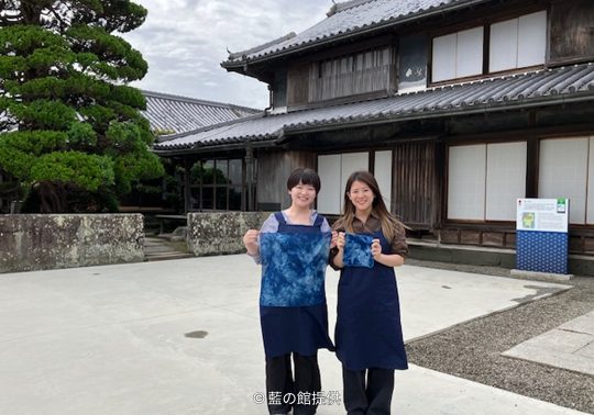 Two women holding newly indigo-dyed cloth pieces standing in the courtyard in front of the traditional wooden Okumura Family Residence.