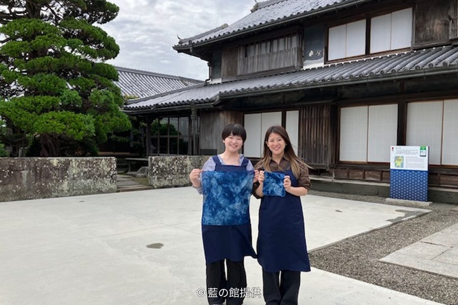 Two women holding newly indigo-dyed cloth pieces standing in the courtyard in front of the traditional wooden Okumura Family Residence.