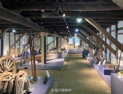 The interior of a traditional wooden storehouse with exposed beams, displaying historical artifacts, tools, and a small boat on pedestals.