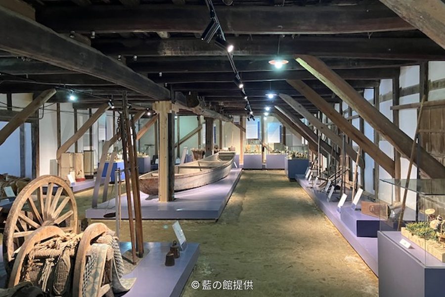 The interior of a traditional wooden storehouse with exposed beams, displaying historical artifacts, tools, and a small boat on pedestals.
