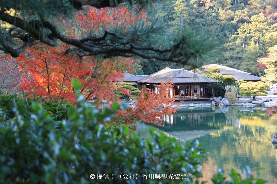 The Kikugetsutei teahouse and pond viewed through the red and orange autumn foliage of foreground trees.