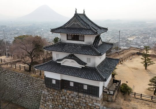Marugame Castle keep perched atop its stone walls, overlooking the city on a misty day, with the conical Mount Iino (Sanuki Fuji) in the distance.