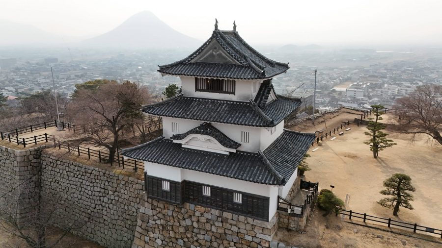 Marugame Castle keep perched atop its stone walls, overlooking the city on a misty day, with the conical Mount Iino (Sanuki Fuji) in the distance.