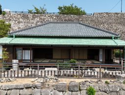 A traditional Japanese-style building (likely a former residence or service building) with a tiled roof, set against a backdrop of a high stone wall.
