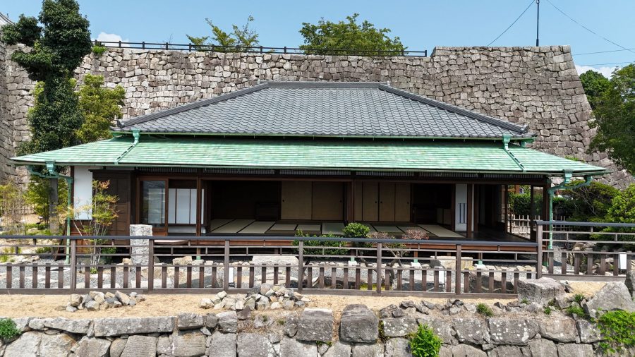 A traditional Japanese-style building (likely a former residence or service building) with a tiled roof, set against a backdrop of a high stone wall.