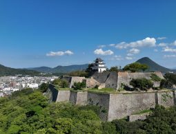 Aerial view of Marugame Castle's towering stone walls and keep on the hill, overlooking the city and surrounding mountains (likely Sanuki Fuji).
