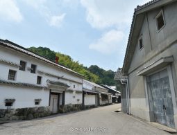 A street in Sakawa Town lined with traditional Japanese buildings featuring white plaster walls, stone bases, and tiled roofs, with a forested hill in the background.