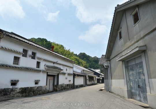 A street in Sakawa Town lined with traditional Japanese buildings featuring white plaster walls, stone bases, and tiled roofs, with a forested hill in the background.
