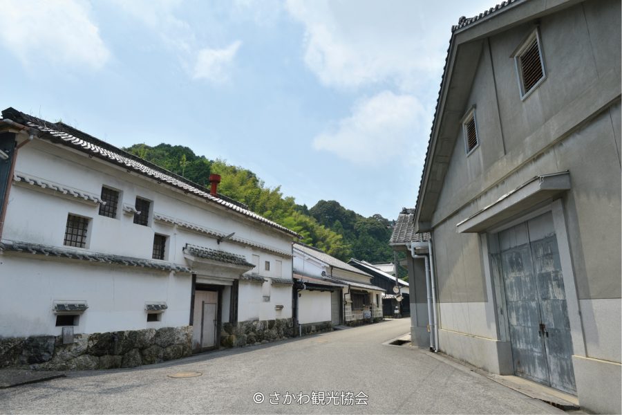 A street in Sakawa Town lined with traditional Japanese buildings featuring white plaster walls, stone bases, and tiled roofs, with a forested hill in the background.