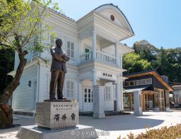 A bronze statue of a man standing in front of a historic white two-story Western-style building with columns and a balcony, under a clear blue sky.