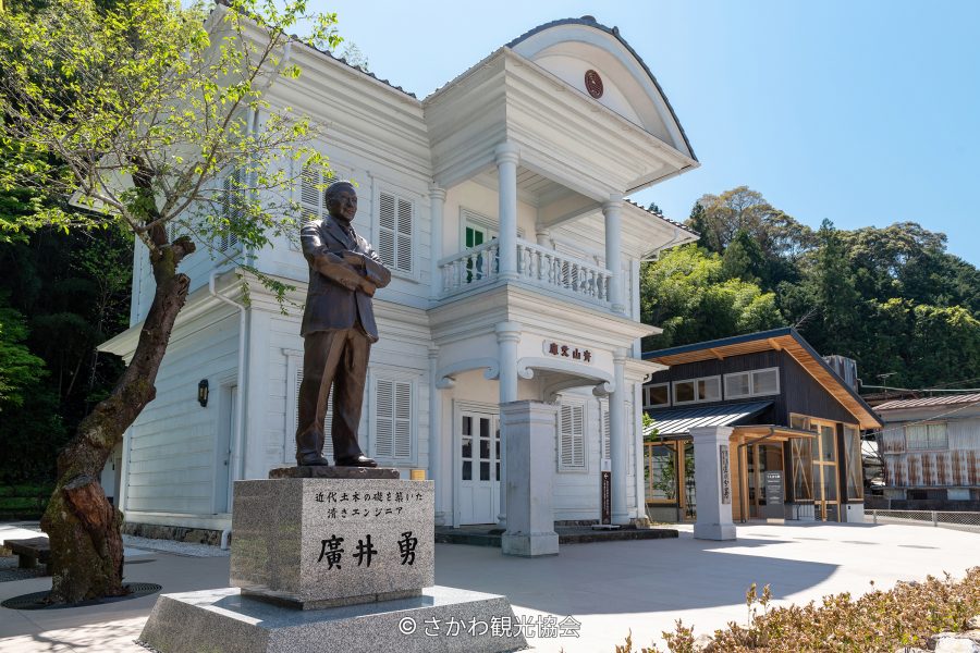 A bronze statue of a man standing in front of a historic white two-story Western-style building with columns and a balcony, under a clear blue sky.