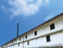A very long, traditional white-walled storehouse with a dark tiled roof and small barred windows, stretching along a quiet street with a tall chimney in the distance.