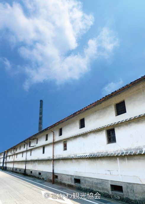 A very long, traditional white-walled storehouse with a dark tiled roof and small barred windows, stretching along a quiet street with a tall chimney in the distance.