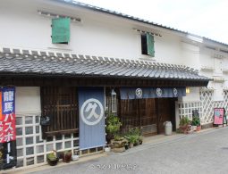 A traditional Japanese merchant house with white walls and a tiled roof, featuring blue curtains (noren) with a white circular logo hanging over the entrance.