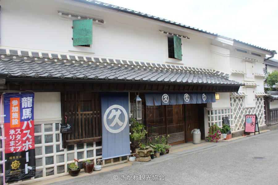 A traditional Japanese merchant house with white walls and a tiled roof, featuring blue curtains (noren) with a white circular logo hanging over the entrance.