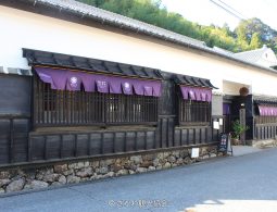 The dark wooden exterior of a traditional Japanese building with purple curtains hanging above the windows, situated along a clean, paved road.