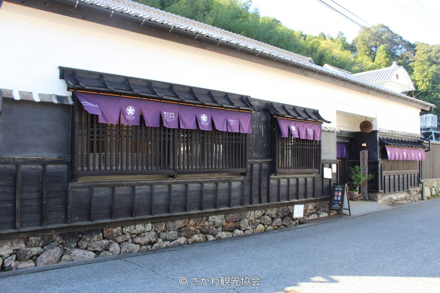 The dark wooden exterior of a traditional Japanese building with purple curtains hanging above the windows, situated along a clean, paved road.
