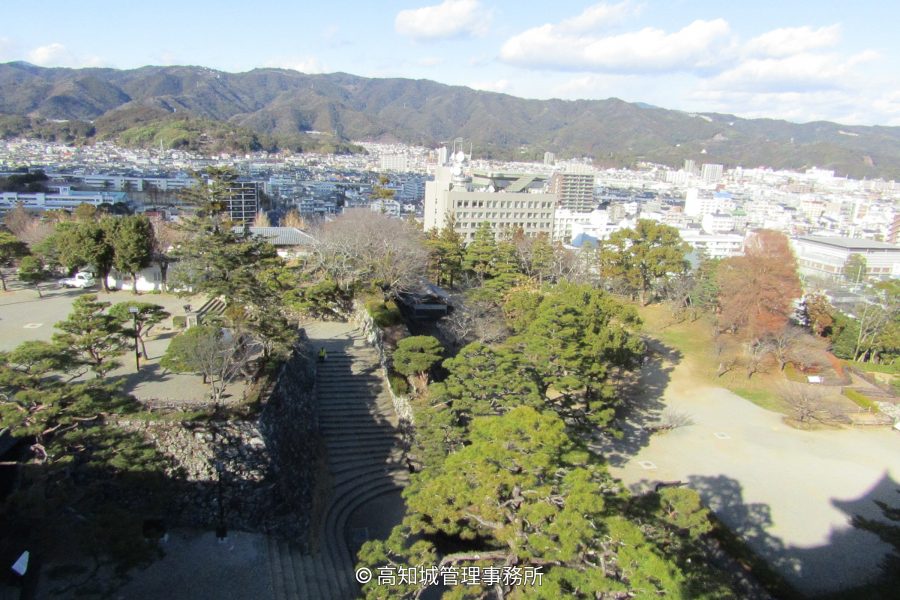 A view from the Kochi Castle walls overlooking the city and surrounding mountains, showing a stone staircase and park area in the foreground.