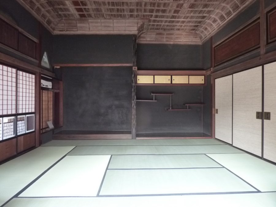 The interior of a traditional Japanese room in a preserved merchant residence, featuring tatami mats, dark wood paneling, and a decorative ceiling.