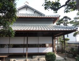A traditional Japanese merchant house (machiya) with a tiled roof, seen through trees in a courtyard.