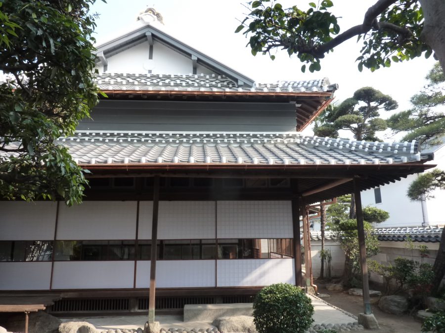 A traditional Japanese merchant house (machiya) with a tiled roof, seen through trees in a courtyard.