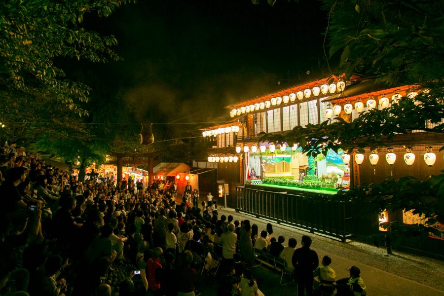 A large crowd of people watching a traditional performance or puppet show on an outdoor stage at night, illuminated by numerous lanterns.