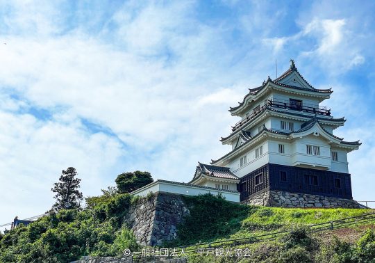 Hirado Castle keep with white walls and black siding, built on a stone base on a green hill under a cloudy blue sky.
