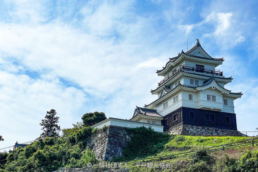 Hirado Castle keep with white walls and black siding, built on a stone base on a green hill under a cloudy blue sky.