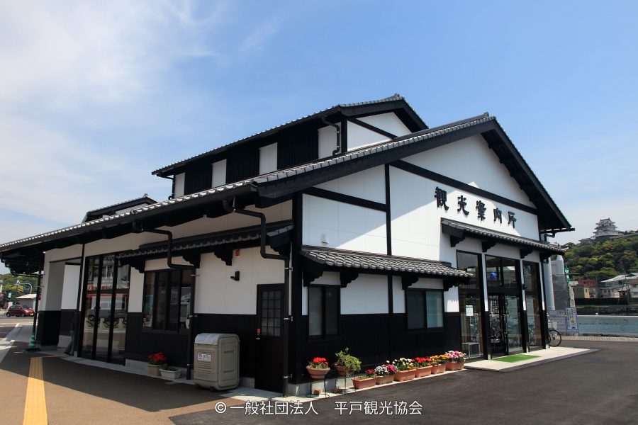 The renovated Hirado Tourist Information Center building, featuring traditional black and white dozo-style architecture, with the castle visible on the hill in the distance.