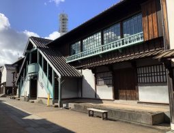 A two-story reconstructed historical building in Dejima, with exterior stairs and a balcony featuring teal-colored railings.