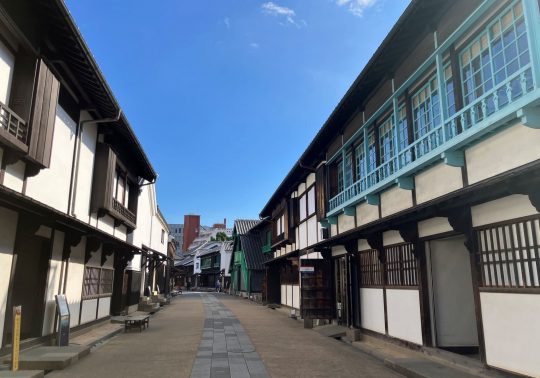 The preserved streetscape inside Dejima, lined with reconstructed traditional buildings featuring white walls and dark wood, with bright blue accents.