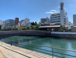 The modern bridge crossing the water channel to the reconstructed Dejima, with traditional buildings and contemporary city architecture in the background.