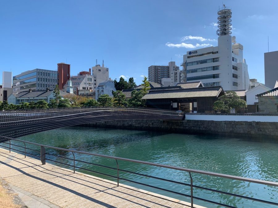 The modern bridge crossing the water channel to the reconstructed Dejima, with traditional buildings and contemporary city architecture in the background.