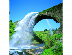 The massive stone arch of Tsujun Bridge aqueduct, dramatically discharging a huge waterfall of water into the valley below on a sunny day.