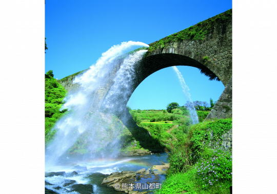 The massive stone arch of Tsujun Bridge aqueduct, dramatically discharging a huge waterfall of water into the valley below on a sunny day.