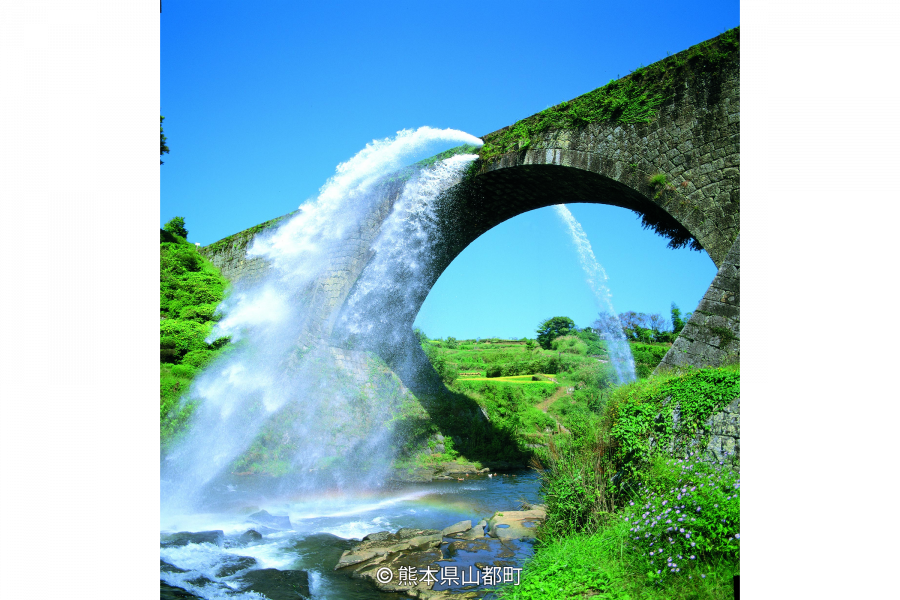 The massive stone arch of Tsujun Bridge aqueduct, dramatically discharging a huge waterfall of water into the valley below on a sunny day.