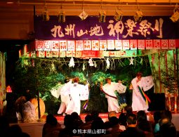 An indoor stage performance of a traditional Japanese Shinto dance (Kagura), featuring dancers in white robes and a banner that reads "Kyushu Mountain Kagura Festival."