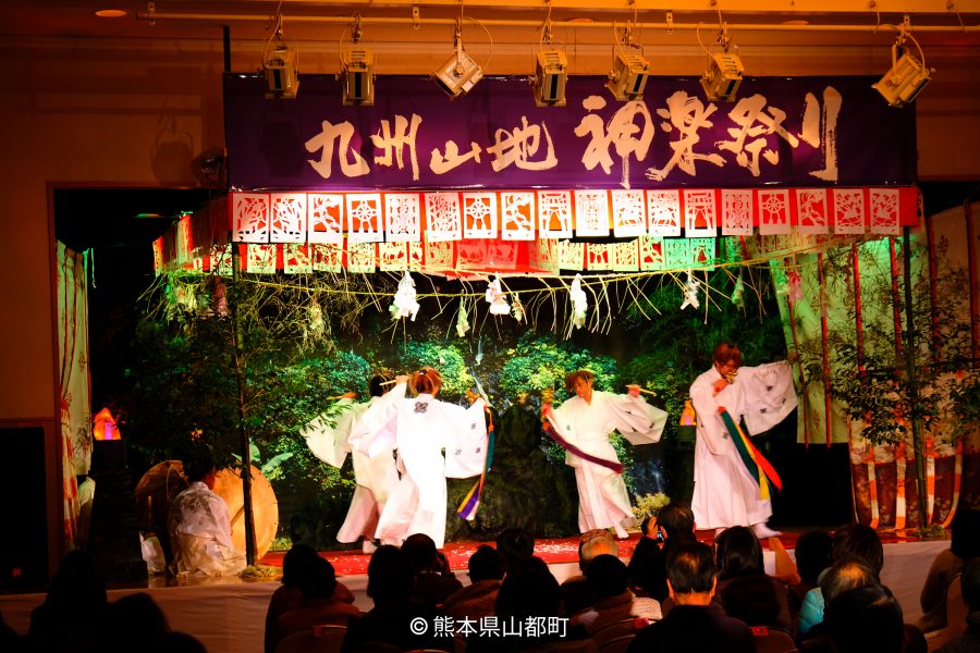 An indoor stage performance of a traditional Japanese Shinto dance (Kagura), featuring dancers in white robes and a banner that reads "Kyushu Mountain Kagura Festival."