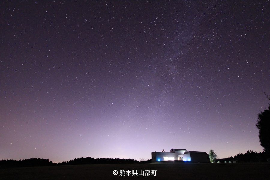 A wide shot of a modern building and distant lights under a dark night sky filled with countless stars.