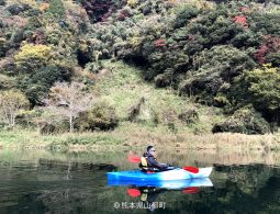 A person kayaking on a calm river surrounded by forested mountains displaying early autumn colors.