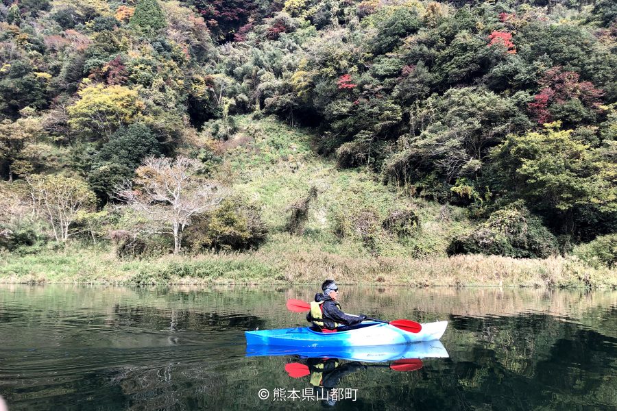 A person kayaking on a calm river surrounded by forested mountains displaying early autumn colors.