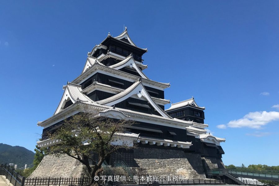 The reconstructed main keep of Kumamoto Castle with black wooden siding and white plaster, rising above its stone walls on a bright, sunny day.