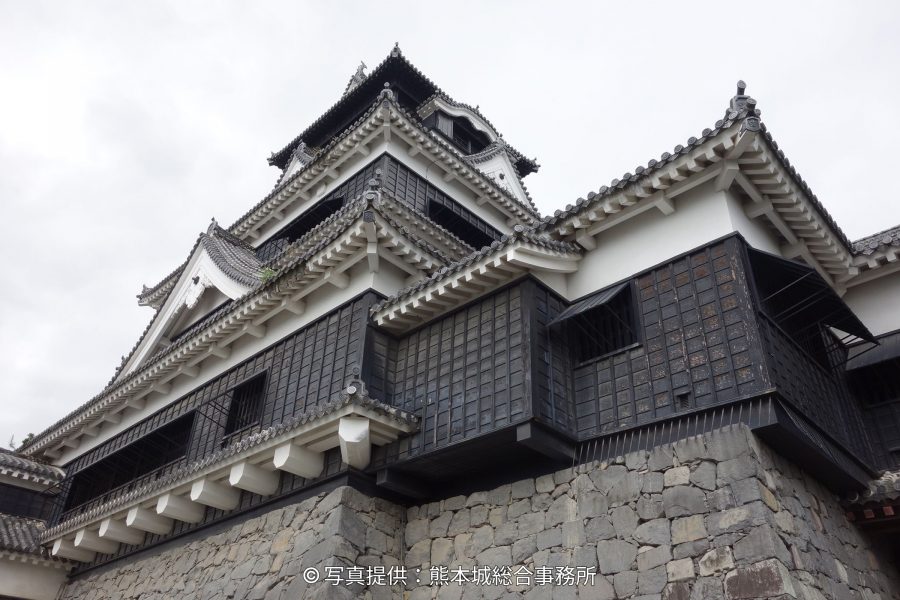 Low-angle view of the large and small keeps of Kumamoto Castle, showing the black, latticed walls and white trim above the rough stone foundation.