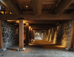 An illuminated stone wall corridor beneath the Kumamoto Castle keep, supported by large wooden beams and pillars.