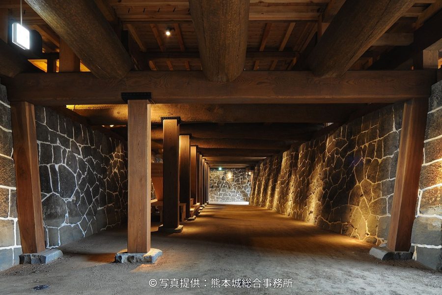An illuminated stone wall corridor beneath the Kumamoto Castle keep, supported by large wooden beams and pillars.