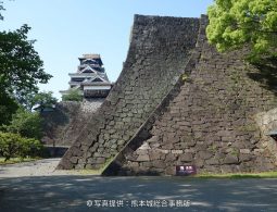 The partially reconstructed black and white castle tower of Kumamoto Castle, visible behind a massive, steeply sloped stone wall (musha-gaeshi) corner.