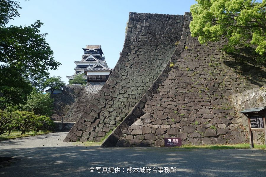 The partially reconstructed black and white castle tower of Kumamoto Castle, visible behind a massive, steeply sloped stone wall (musha-gaeshi) corner.