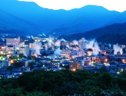 Panoramic view of Beppu city at dusk, showing numerous plumes of hot spring steam rising above the illuminated buildings against a deep blue mountain backdrop.
