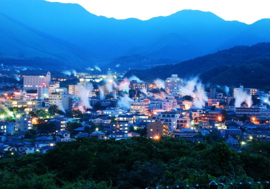 Panoramic view of Beppu city at dusk, showing numerous plumes of hot spring steam rising above the illuminated buildings against a deep blue mountain backdrop.